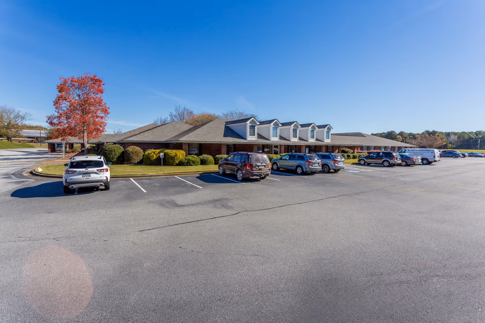 Single-story brick nursing facility with dormer windows, a large parking lot with parked cars, and a red-leaved tree under a clear blue sky.