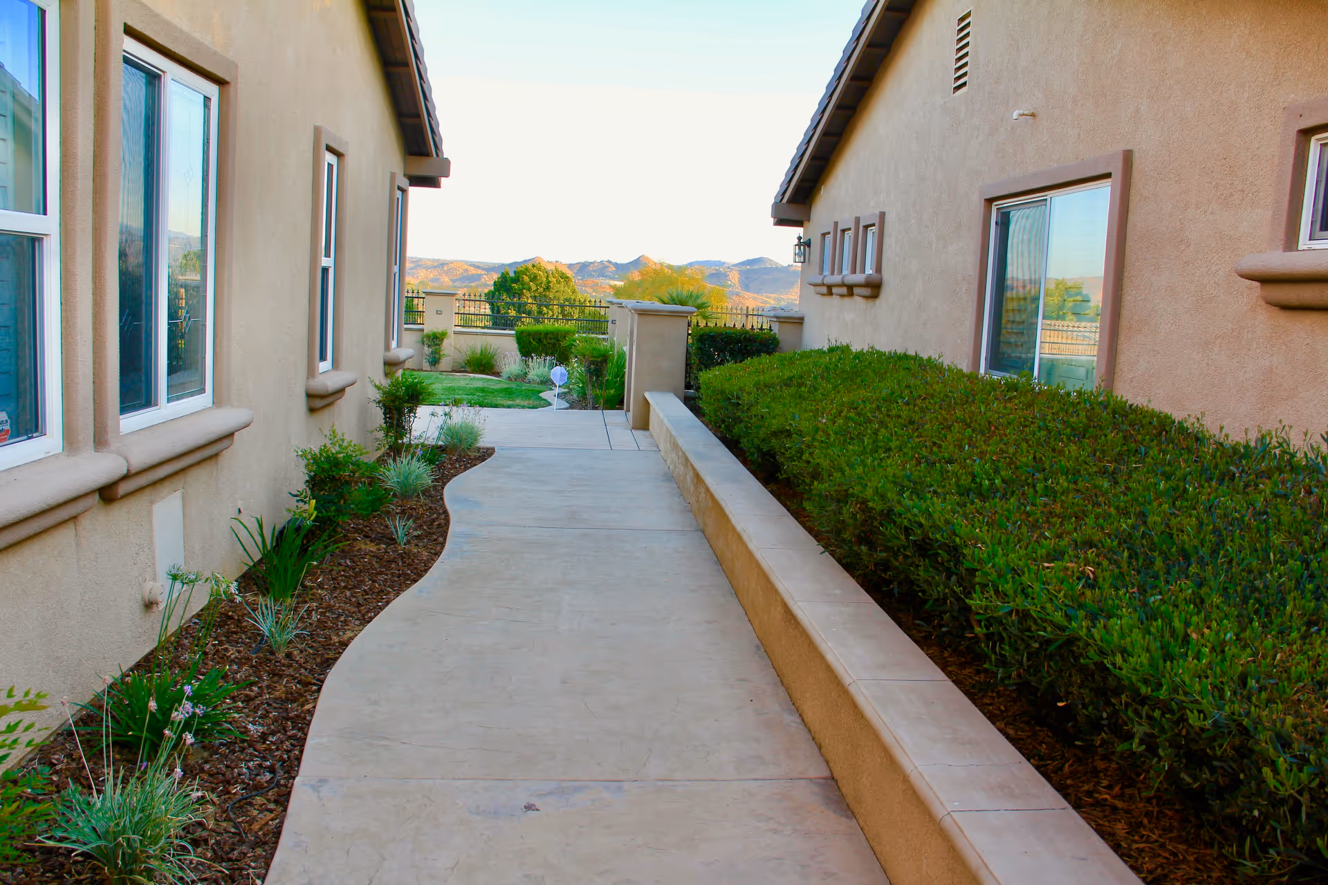 Paved walkway between two beige stucco buildings with landscaped plantings and hills visible in the background.