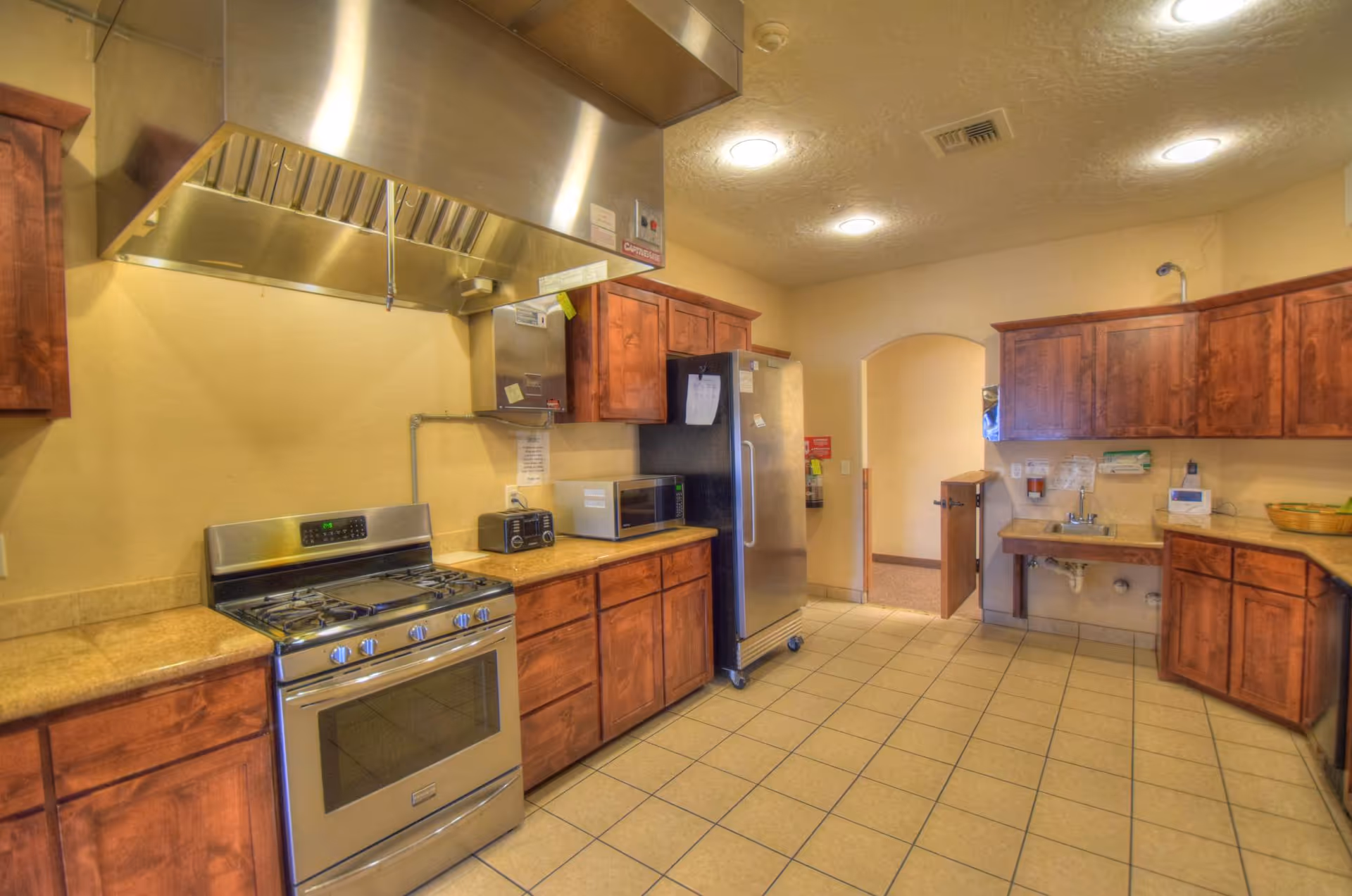 A spacious kitchen with tiled floor, wooden cabinets, a stainless steel stove with an overhead vent hood, a refrigerator, microwave, toaster, and a sink area with a basket on the counter. The kitchen has warm lighting and an arched doorway leading to another room.
