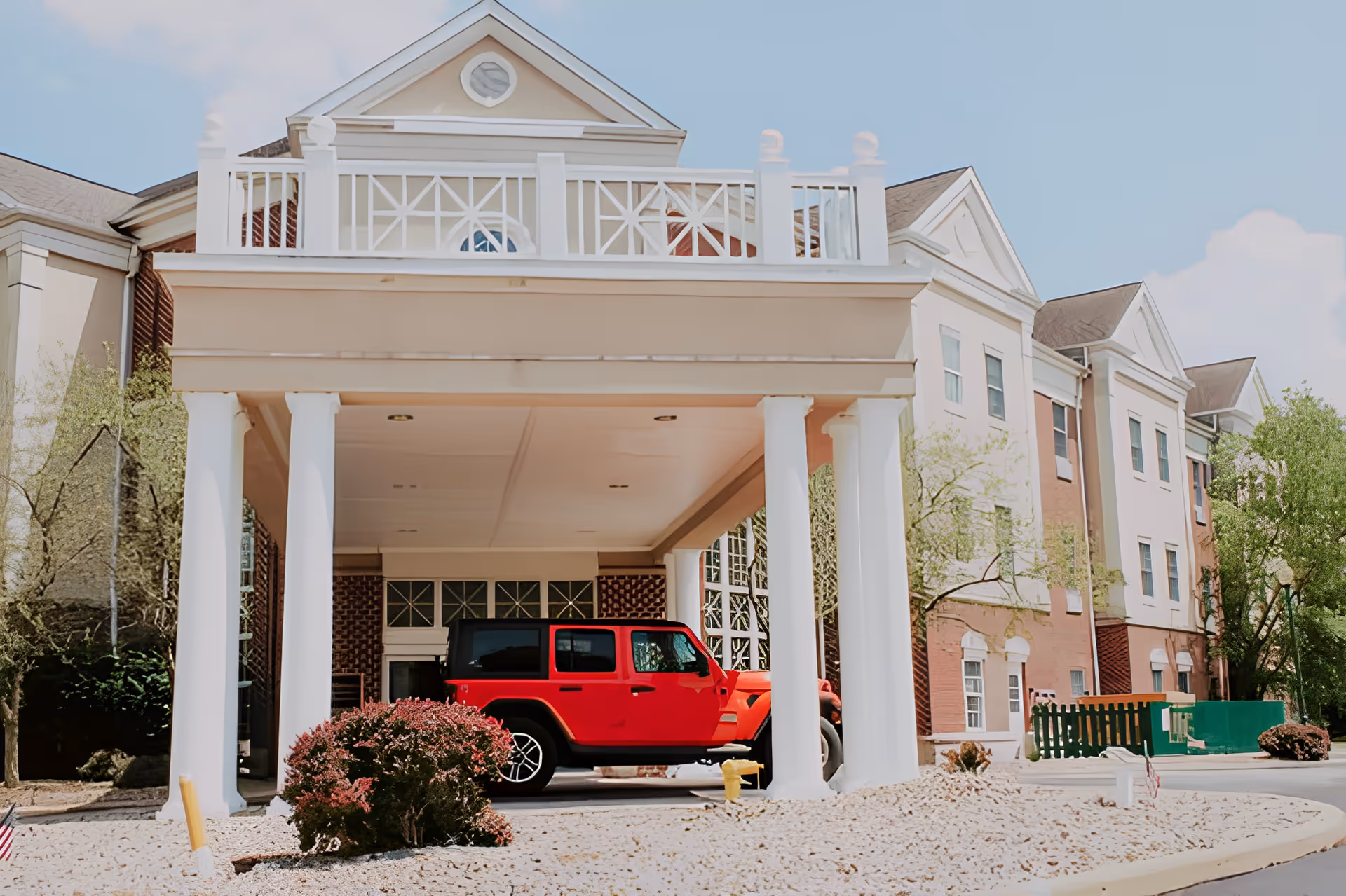 Front entrance of a senior living facility with a covered drop-off area supported by white columns. A red SUV is parked under the covered area. The building has a brick and light-colored facade with multiple windows and a balcony above the entrance. Trees and bushes surround the area under a clear blue sky.