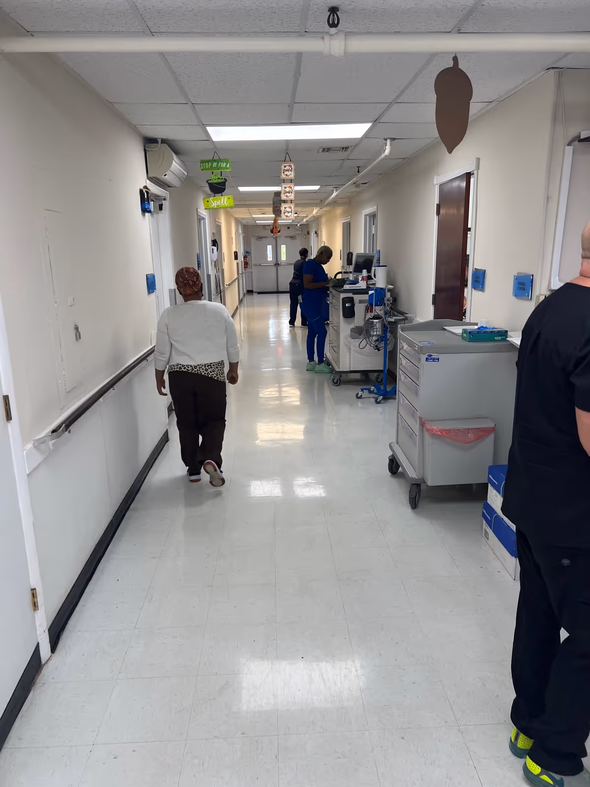 A long, clean hallway in Pines Nursing Home with white walls and a tiled floor. Several people are present, including a person walking away from the camera and two staff members working near medical carts along the right side. The ceiling has fluorescent lights and some hanging decorations.