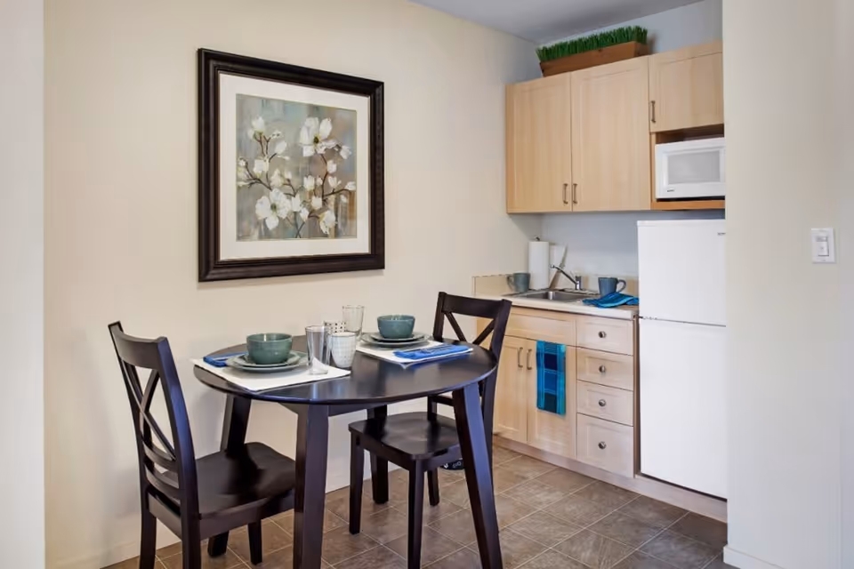 Small kitchen area with light wood cabinets, a white refrigerator, microwave, and a sink. In front of the kitchen is a round dark wood table set for two with green bowls, plates, glasses, and blue napkins. A framed floral painting hangs on the wall above the table.