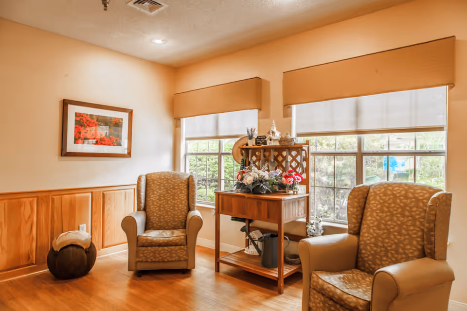 A cozy sitting area with two patterned armchairs placed on a wooden floor. Between the chairs is a wooden side table decorated with artificial flowers and small decorative items. Two large windows with beige valances and white roller shades let in natural light, and a framed picture with red flowers hangs on the wall. The room has light-colored walls with wooden wainscoting.