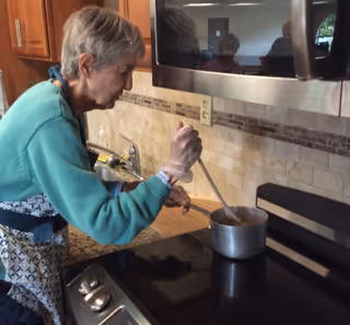 An elderly woman stirring food in a pot on a stove in a kitchen with wooden cabinets and a tiled backsplash.