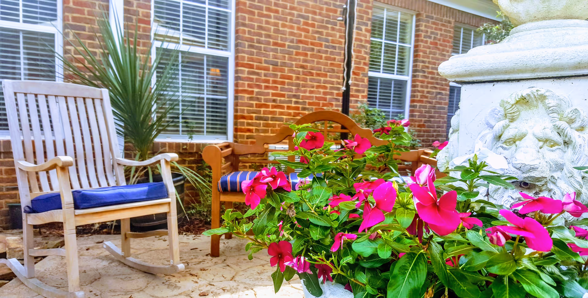 Outdoor patio area with a wooden rocking chair with a blue cushion, a wooden bench with a striped cushion, vibrant pink flowers in the foreground, and a decorative stone pillar with a lion's face. The background shows a brick building with white-framed windows and some greenery.