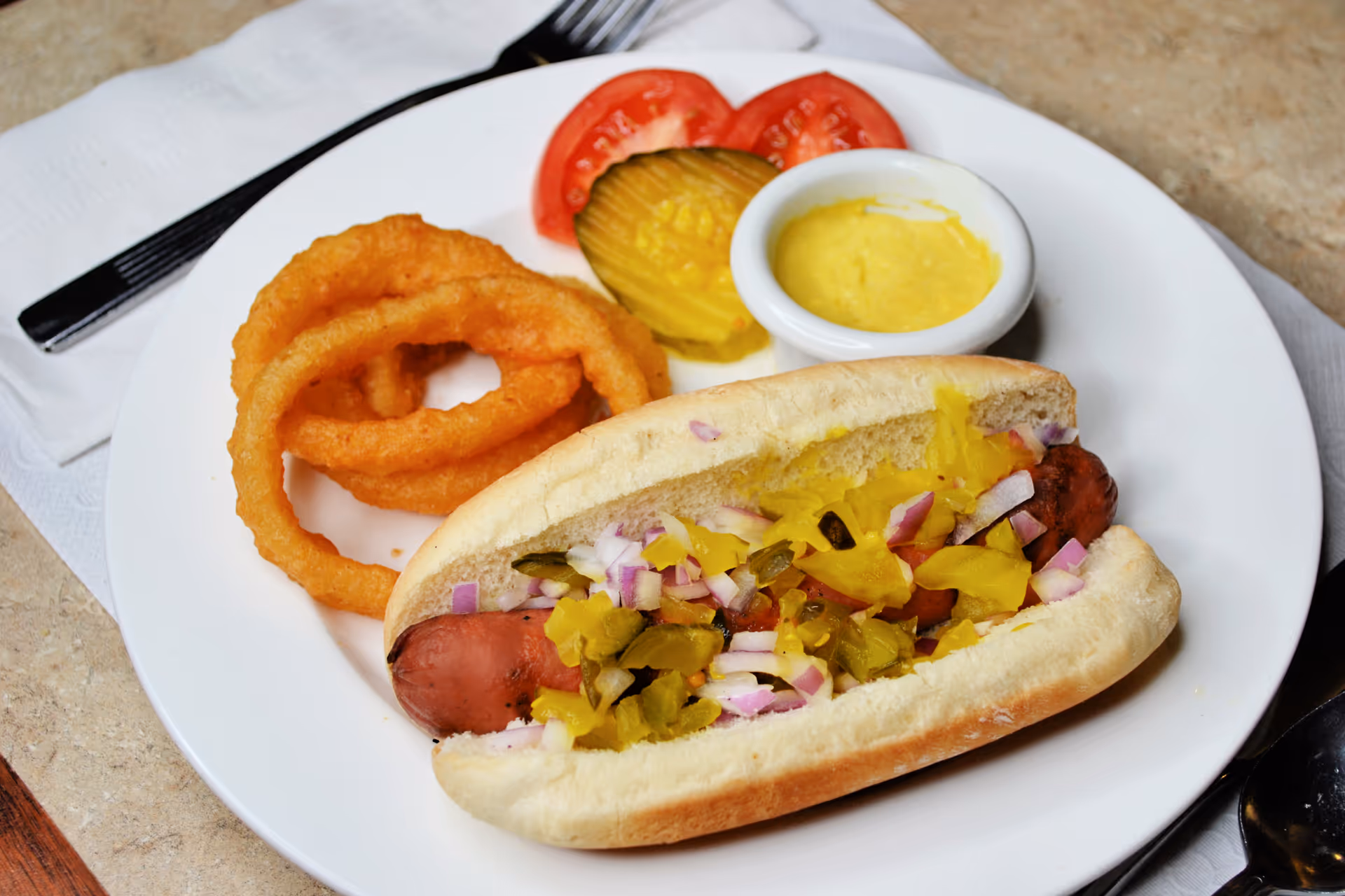 A plate with a hot dog topped with chopped onions and relish, served with three onion rings, two slices of tomato, two pickle slices, and a small cup of yellow mustard. A fork and knife are placed on a white napkin beside the plate.