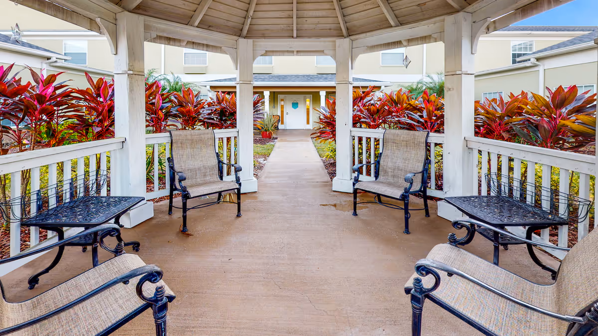 Covered gazebo seating area with chairs and tables leading along a walkway to a building entrance, flanked by red foliage.
