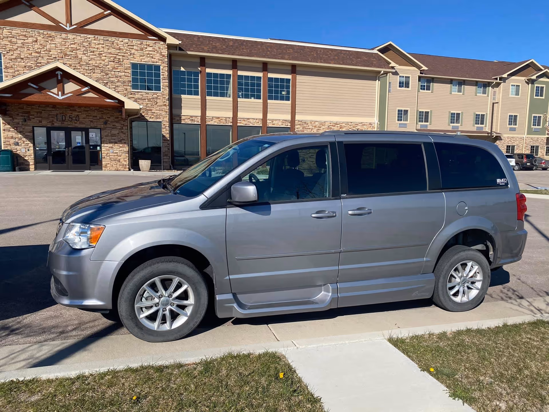 A silver minivan parked on a driveway in front of a large assisted living facility building with stone and beige siding exterior under a clear blue sky.