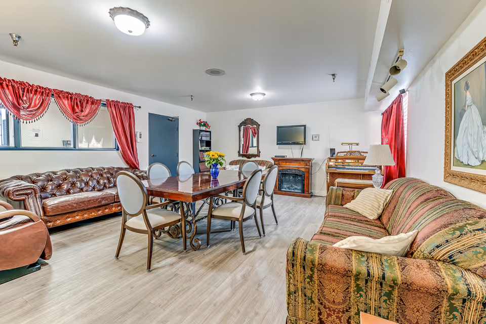 A spacious living room in an assisted living facility featuring a large wooden dining table with six chairs, a brown leather tufted sofa, a patterned fabric sofa with pillows, a small electric fireplace with a mounted TV above it, a mirror on the wall, a wooden organ, and red curtains on the windows. The room has light-colored wood flooring and white walls.
