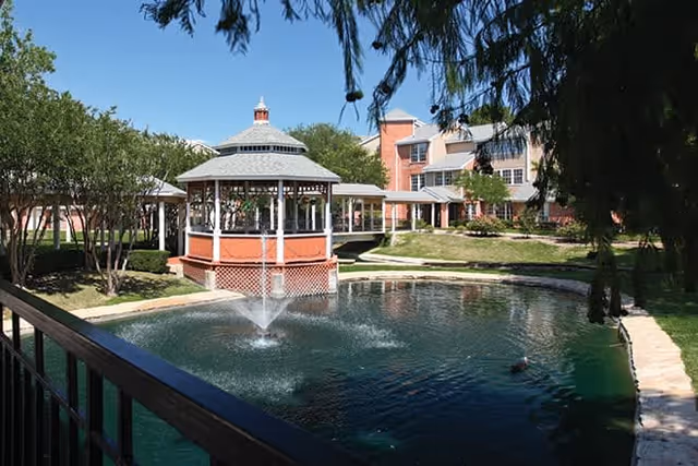 Outdoor view of a senior living facility featuring a gazebo over a pond with a water fountain, surrounded by trees and a multi-story building in the background under a clear blue sky.