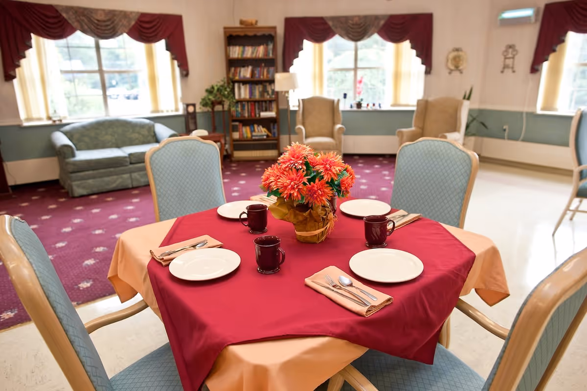 Dining table set for four with a floral centerpiece in a bright senior living common room with upholstered chairs, sofas, and bookshelves.
