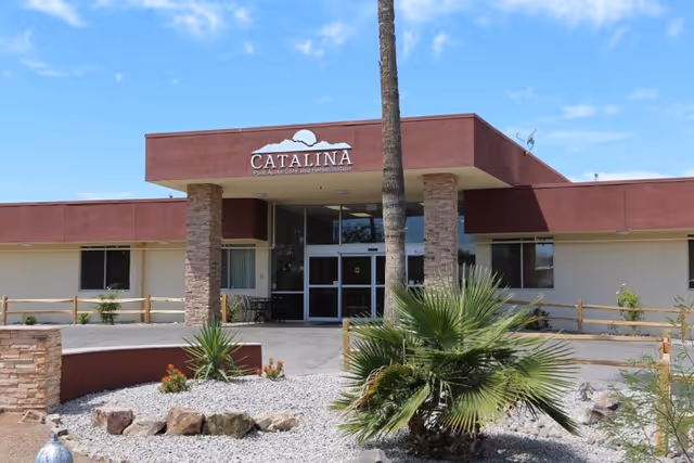 Front exterior view of Catalina Post Acute and Rehabilitation facility with a red and beige building, stone pillars at the entrance, a palm tree, desert landscaping with rocks and plants, and a clear blue sky.