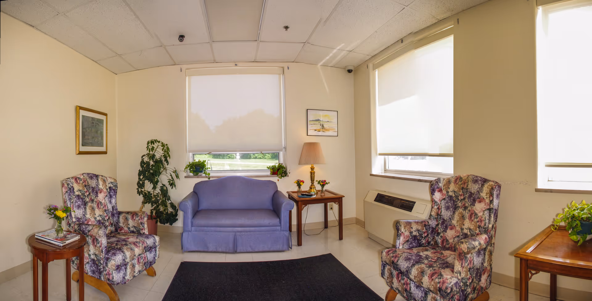 A small sitting area with two floral upholstered armchairs and a purple loveseat arranged around a black rug. There are two wooden side tables, one with a lamp and small flower vases, and the other with a small plant and magazines. The room has cream-colored walls, two windows with white roller shades, and framed artwork on the walls.