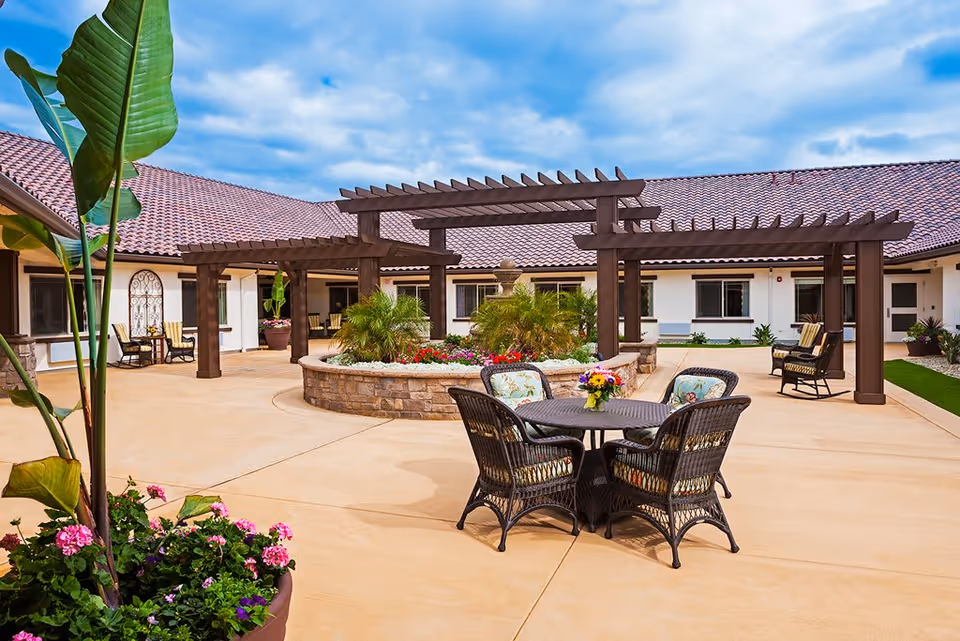 Outdoor courtyard area with a round table and four wicker chairs with floral cushions. The courtyard features a central raised planter with green plants and flowers, surrounded by wooden pergolas. The building has a tiled roof and white walls with windows and doors opening into the courtyard. Potted plants and rocking chairs are also visible around the space under a partly cloudy sky.