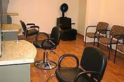 A small salon-style interior with several black styling chairs, a countertop with mirrors, and waiting chairs along the wall.