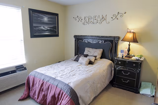 A neatly made single bed with an ornate headboard, 'DREAM' wall art above, a bedside table with lamp, and a window with blinds in a small bedroom.