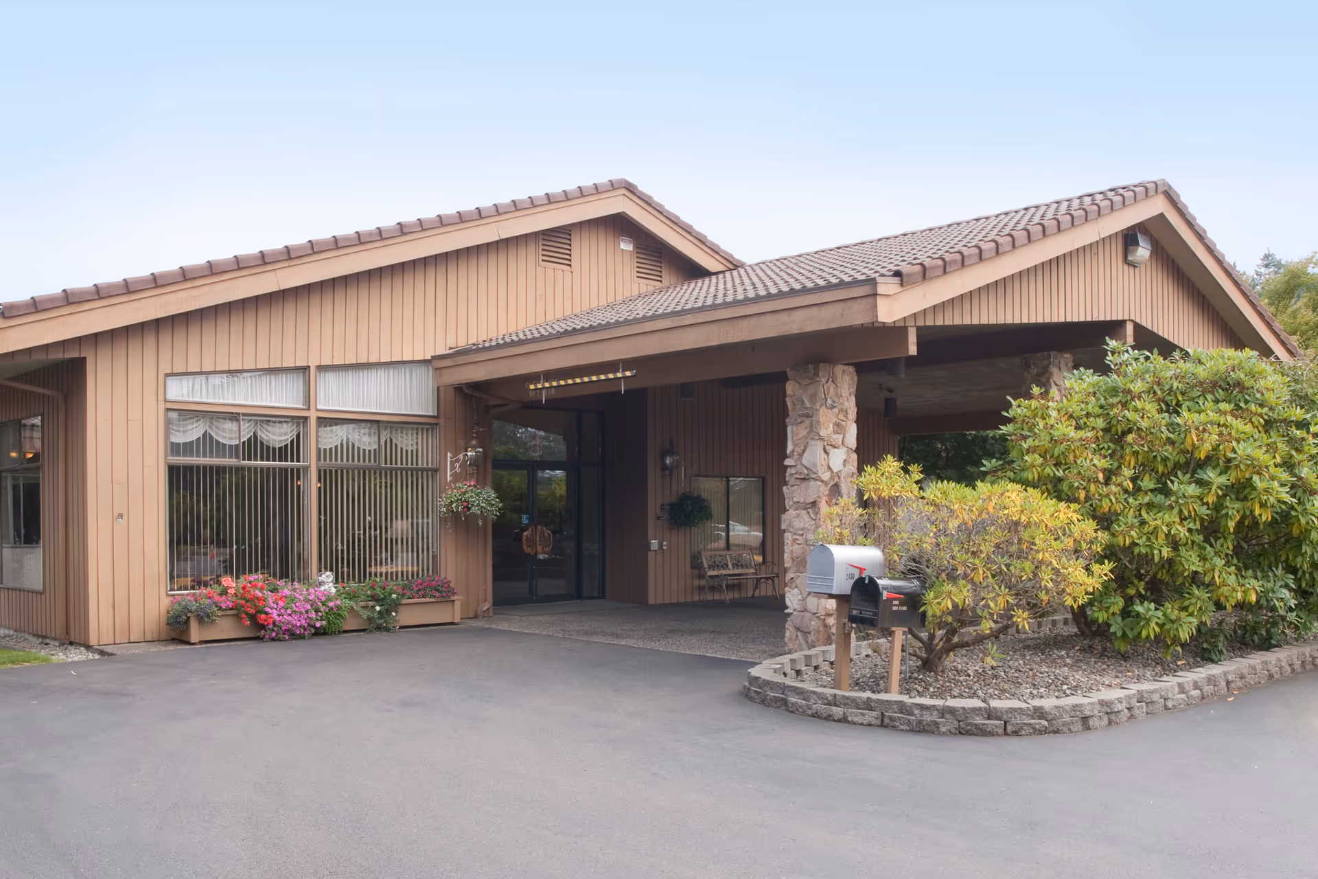 Exterior view of a single-story building with a sloped roof and large windows. The entrance is covered by a porch supported by stone pillars. There are flower beds with colorful flowers and shrubs near the entrance, along with two mailboxes on a small landscaped area beside the driveway.