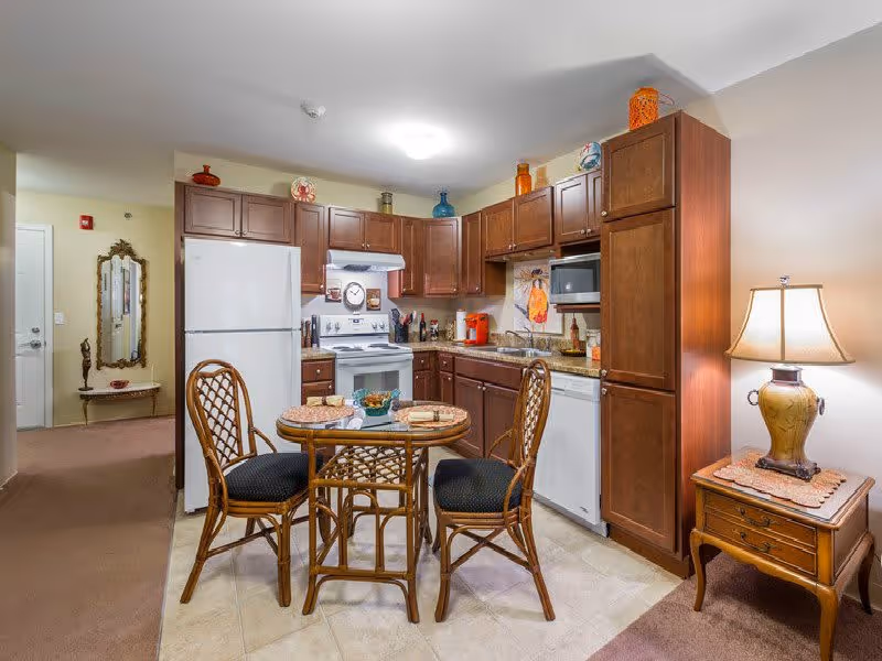 A cozy kitchen area in a retirement community featuring wooden cabinets, a white refrigerator, stove, microwave, and dishwasher. In front of the kitchen is a small round glass-top table with three wicker chairs. To the right, there is a wooden side table with a decorative lamp. The kitchen is well-lit with ceiling lights and decorated with colorful vases and artwork.
