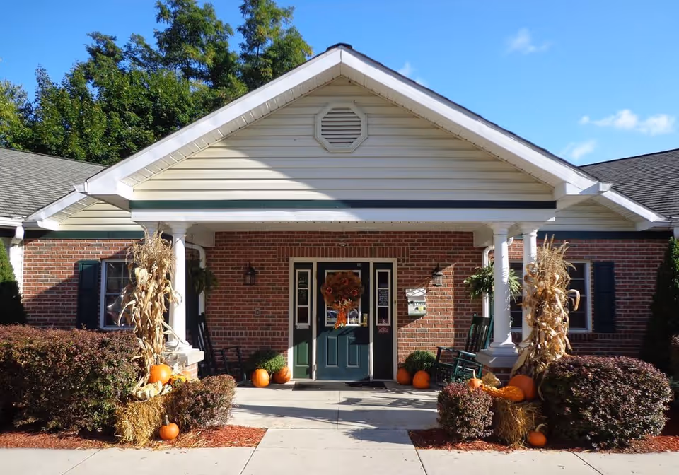 Front entrance of a brick building with a green door decorated with a fall wreath. The entrance is flanked by white columns and rocking chairs. There are autumn decorations including pumpkins, hay bales, and dried corn stalks on either side of the walkway leading to the door. Green bushes and trees surround the entrance under a clear blue sky.