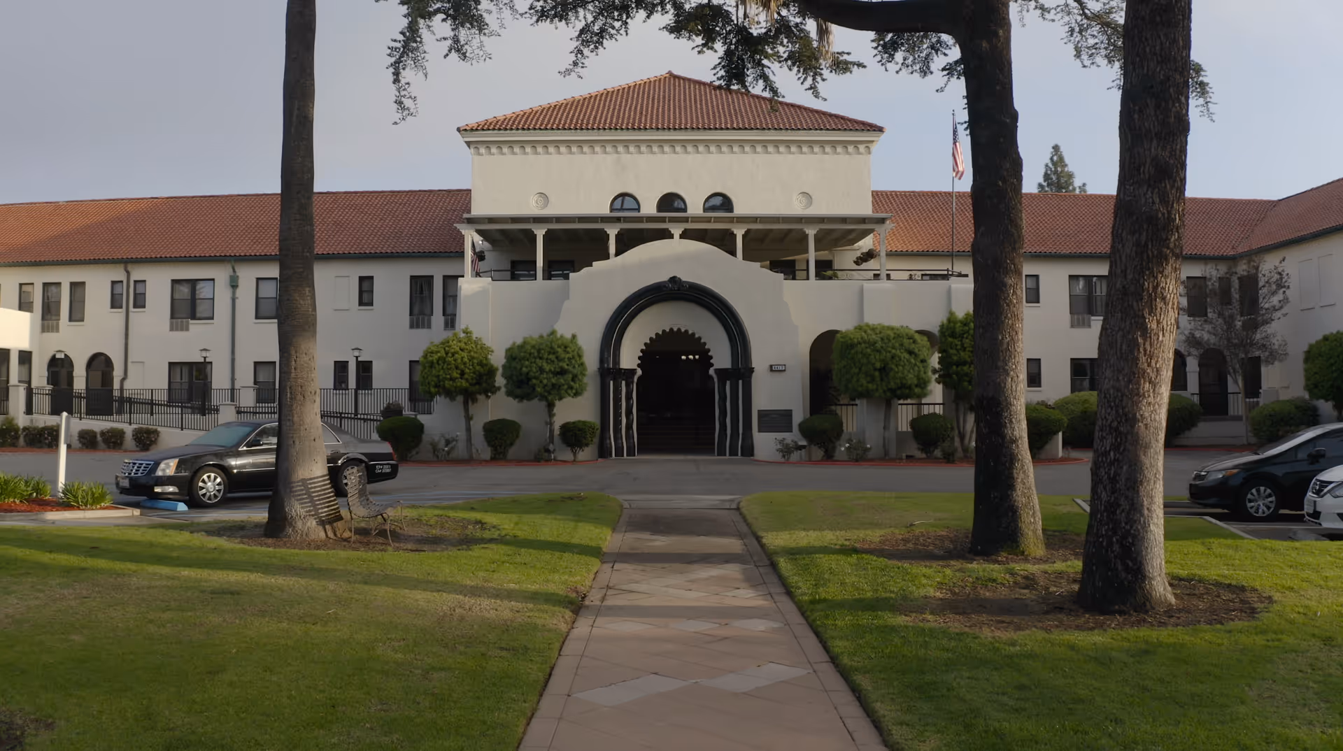 Front exterior view of the California Mission Inn building with a tiled roof, arched entrance, and manicured lawn with trees and benches in the foreground. Several cars are parked on either side of the entrance driveway.