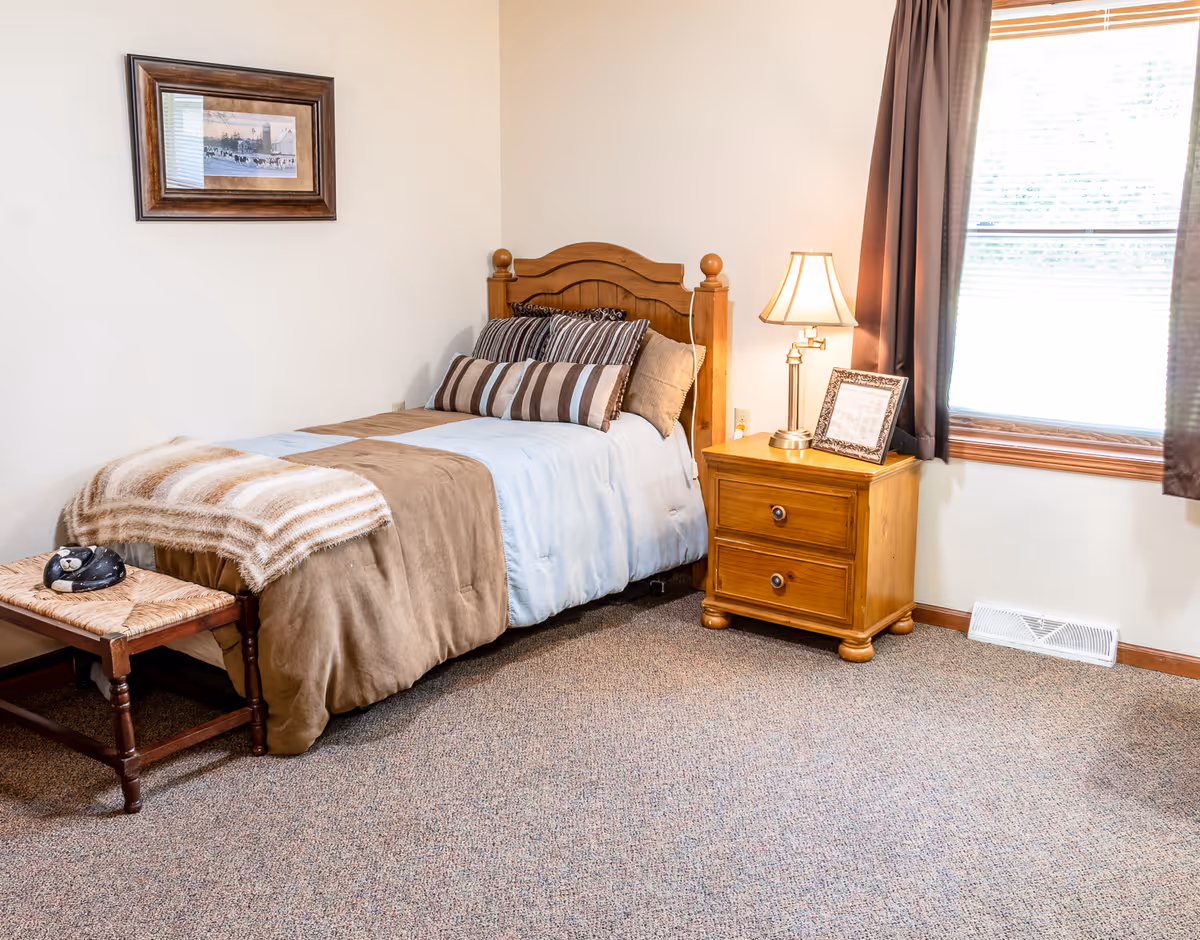 A cozy single bedroom with a wooden headboard bed, striped pillows and blanket, a wooden nightstand with lamp and framed photo, and a window with curtains.
