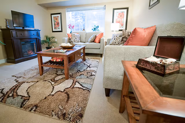 A cozy living room with beige sofas adorned with patterned and solid orange pillows, a wooden coffee table on a patterned area rug, a flat-screen TV on a wooden stand with an electric fireplace, and framed floral artwork on the walls.