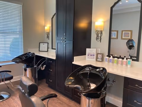 Interior salon area with two shampoo sinks and chairs, mirrors, dark cabinets, and hair products on the counter.