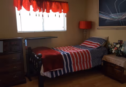 Single bedroom in a senior facility with patriotic bedding and a window with red curtains.