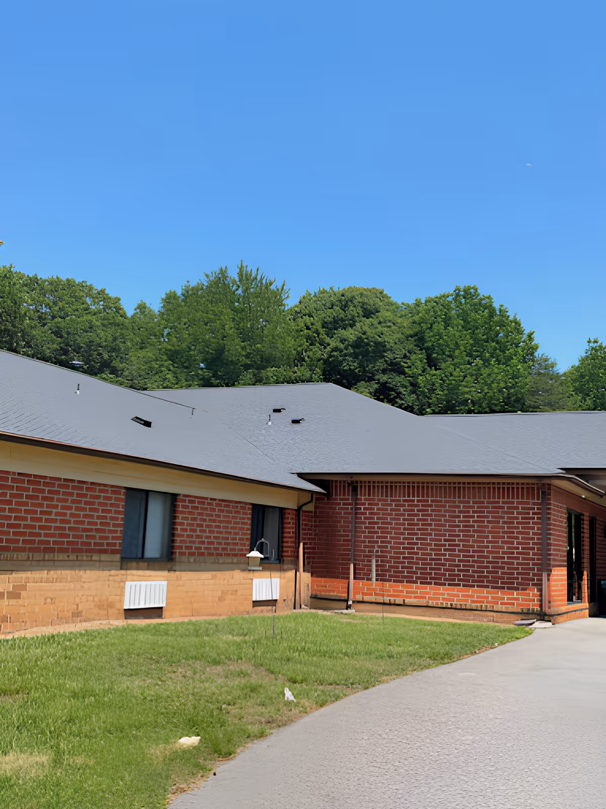 Exterior view of a single-story brick building with a gray roof, surrounded by green grass and trees under a clear blue sky.