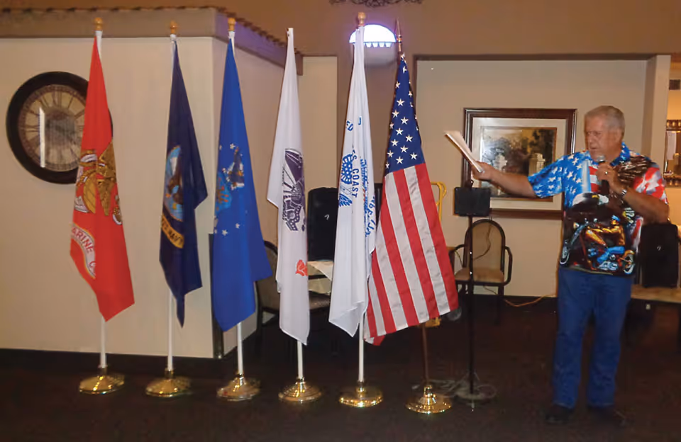 An elderly man wearing a patriotic shirt stands indoors next to six flags on stands, including the American flag and various military service flags. The room has beige walls, a framed picture, and a large clock on the wall.