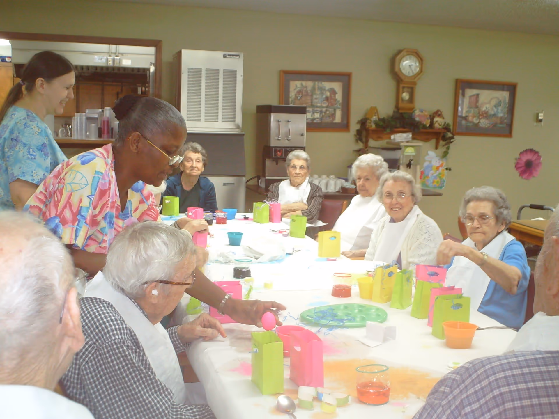 A group of elderly people sitting around a long table engaged in an activity involving colorful paper bags and cups, with two caregivers assisting them in a well-lit room decorated with framed pictures and a clock on the wall.