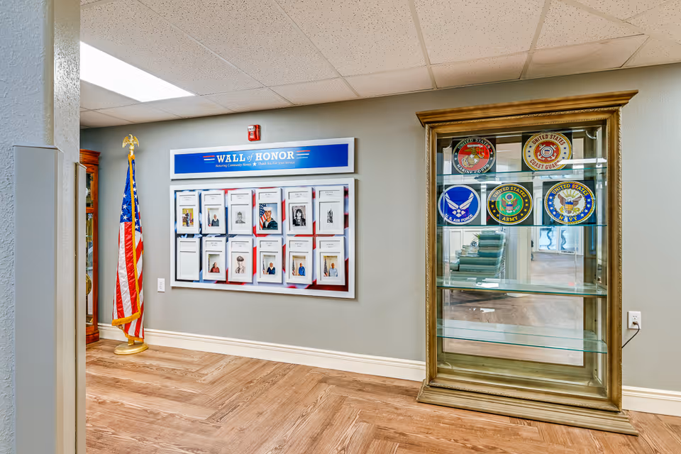Interior view of a hallway in a senior living facility featuring a Wall of Honor display with photos and names of veterans, an American flag on a stand, and a glass display cabinet with emblems of different United States military branches.