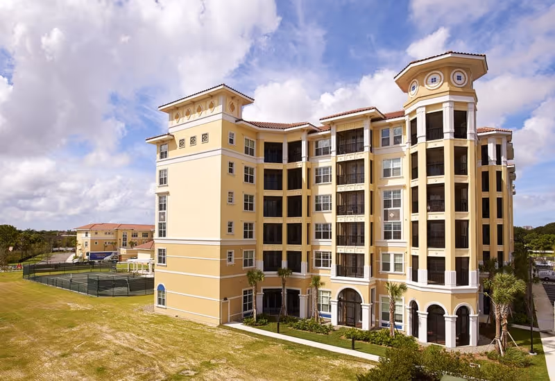 Exterior view of a multi-story yellow building with arched windows and balconies, surrounded by a grassy area and a tennis court, under a partly cloudy sky.