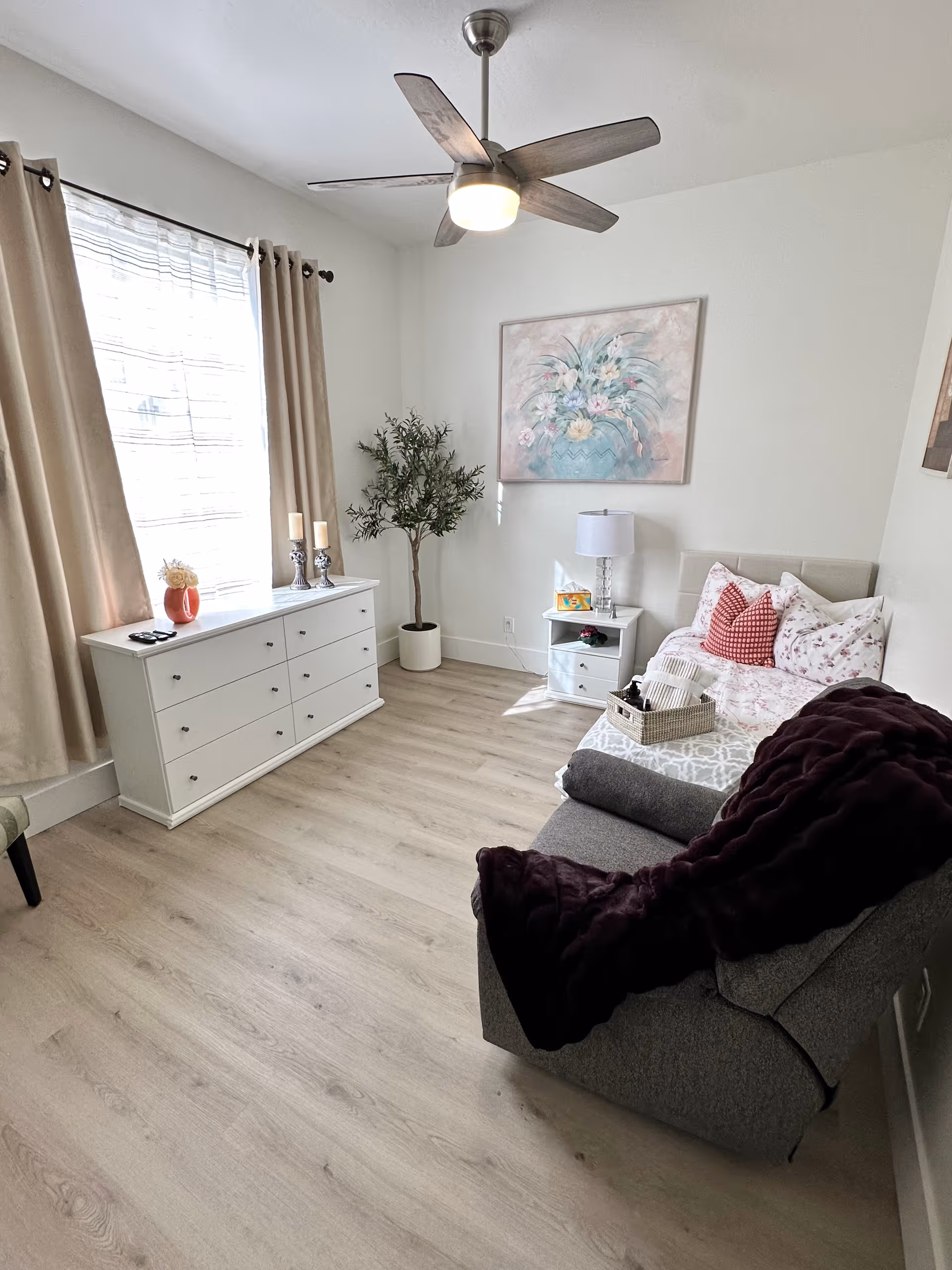 A bright and cozy bedroom with light wood flooring, a white dresser under a window with beige curtains, a small white nightstand with a lamp, a bed with floral and red pillows, a gray armchair with a dark purple throw blanket, a ceiling fan with light, a potted plant in the corner, and a floral painting on the wall.
