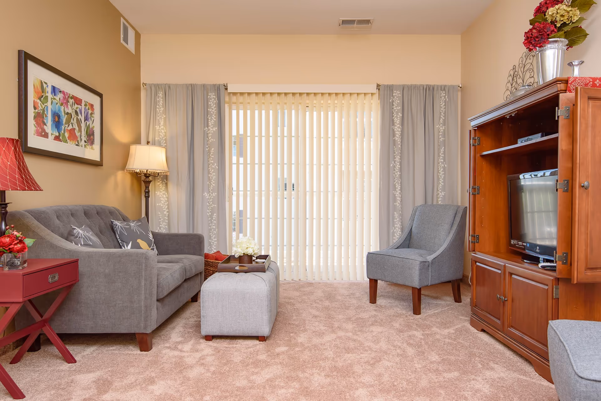 Cozy living room with a gray sofa and chair, ottoman, wooden TV cabinet, side tables, lamps, and vertical blinds covering a sliding door.