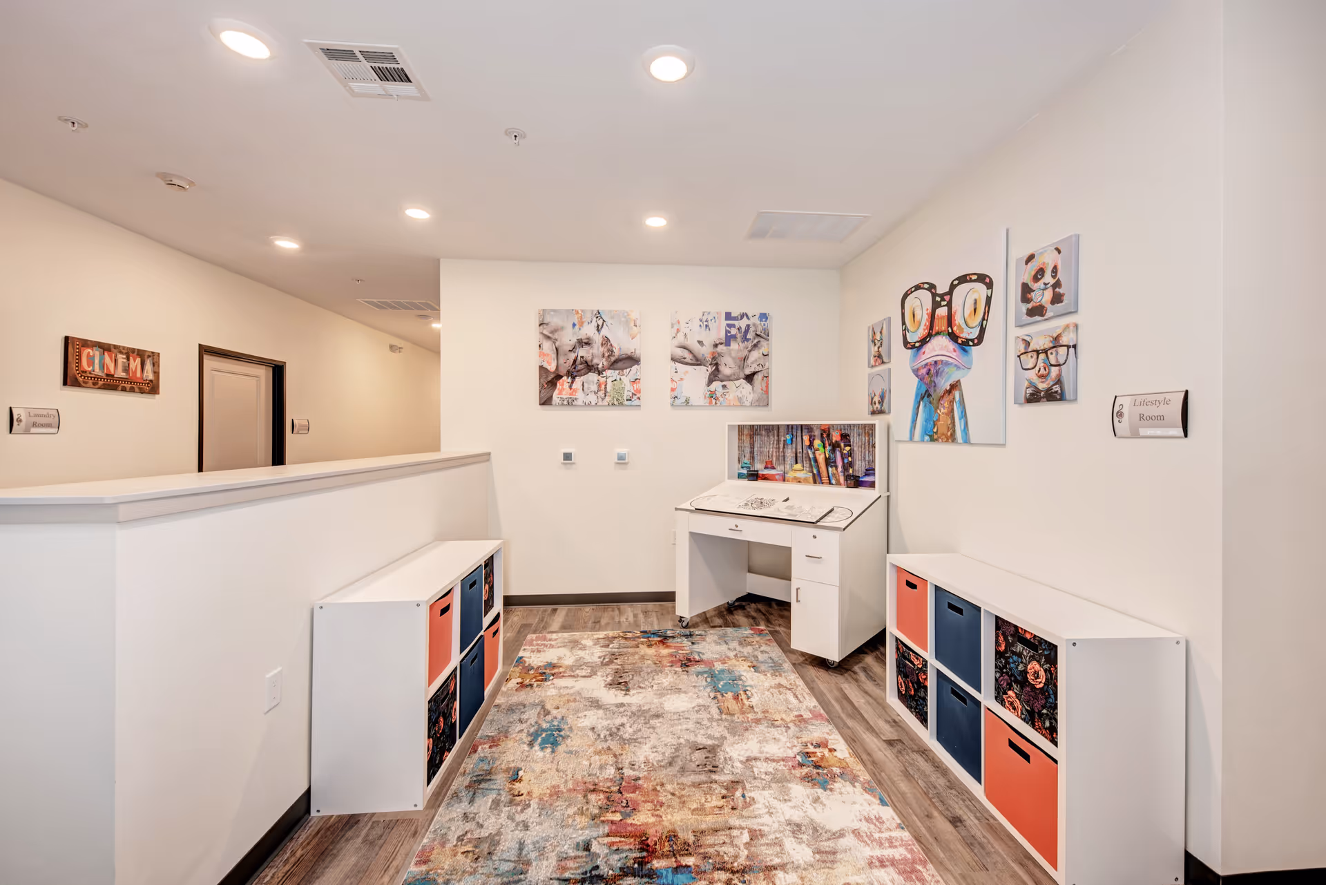 A bright and colorful interior room with white walls and wood flooring. The room features two white storage units with colorful bins on either side of a patterned area rug. At the far end, there is a white desk with art supplies and colorful artwork on the walls, including animal portraits wearing glasses. Signs on the walls indicate this is a Lifestyle Room and a nearby Cinema room.