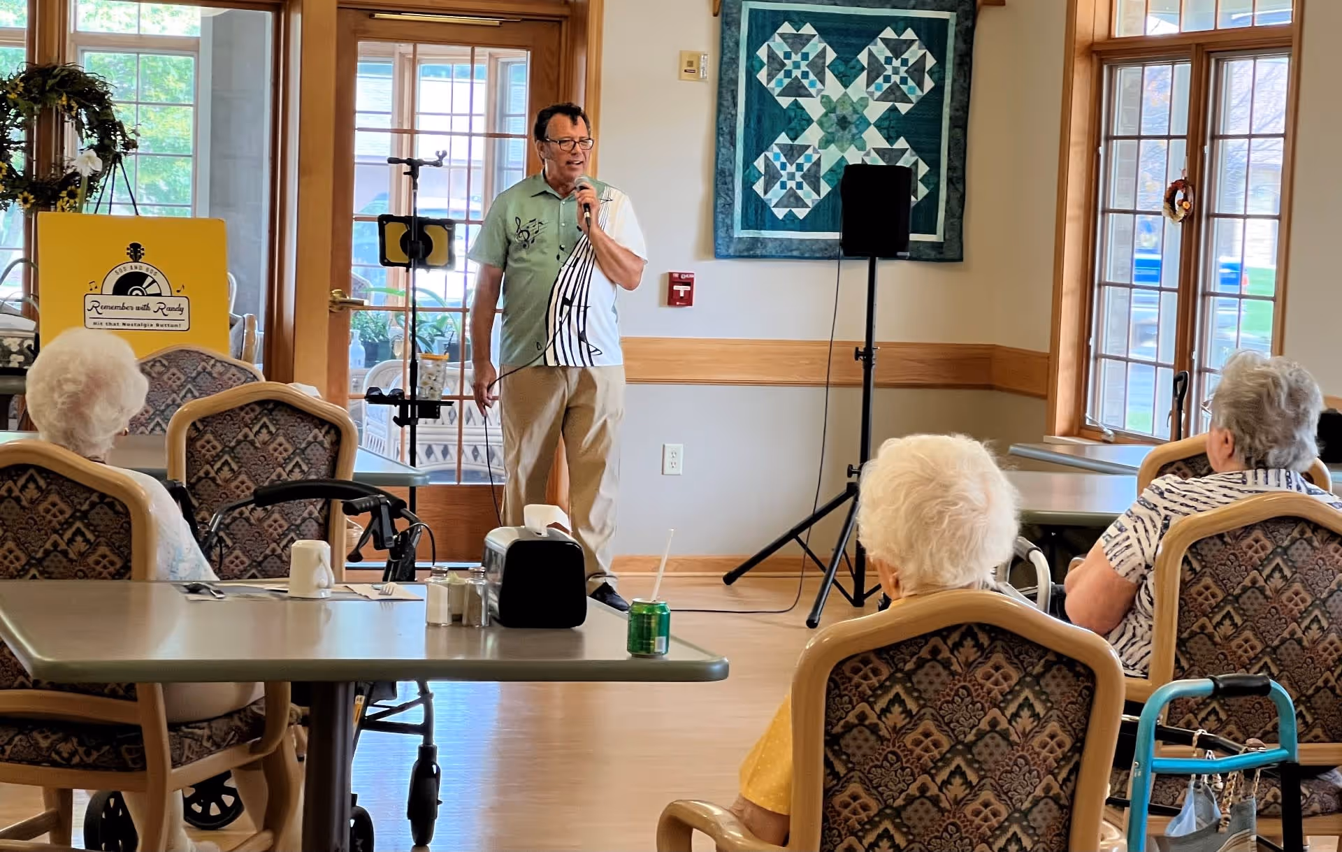 A man holding a microphone is performing or speaking in a room with several elderly people seated and watching him. The room has large windows, a quilt hanging on the wall, and a sign that reads 'Remember with Randy'. Some of the elderly attendees have walkers beside them.