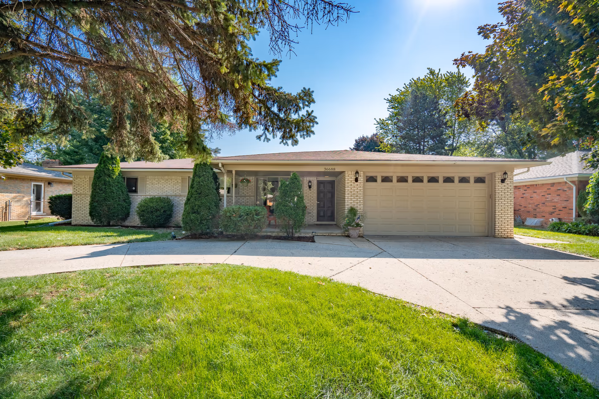 Front exterior view of a single-story brick house with a two-car garage, a driveway, and a well-maintained lawn with trees and shrubs under a clear blue sky.