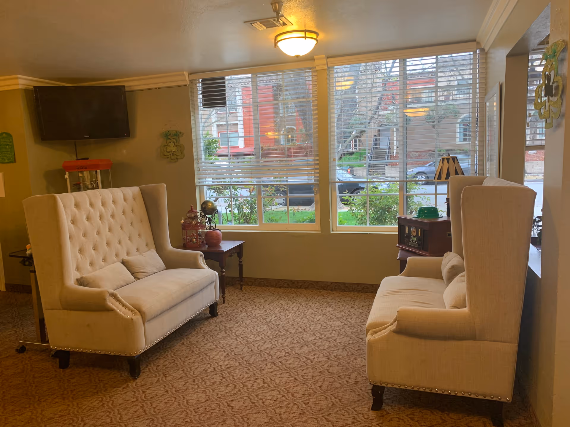 A cozy sitting area with two beige tufted armchairs facing each other, a small wooden side table with decorative items between them, a flat-screen TV mounted on the wall, and large windows with blinds letting in natural light. The room has a patterned carpet and soft lighting from a ceiling fixture.