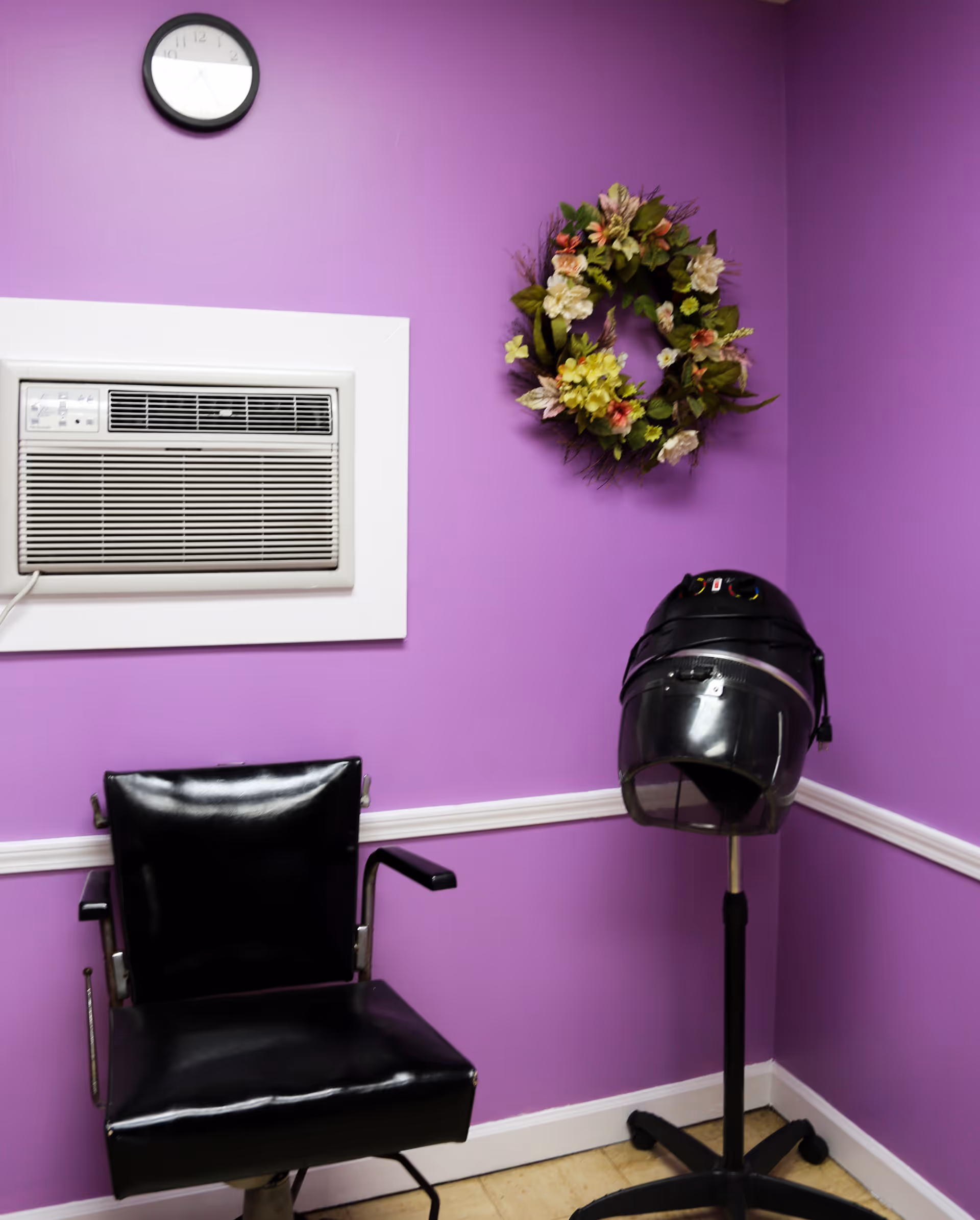A small room with purple walls featuring a black salon chair, a standing hair dryer, a floral wreath on the wall, a wall-mounted air conditioner, and a clock above the air conditioner.