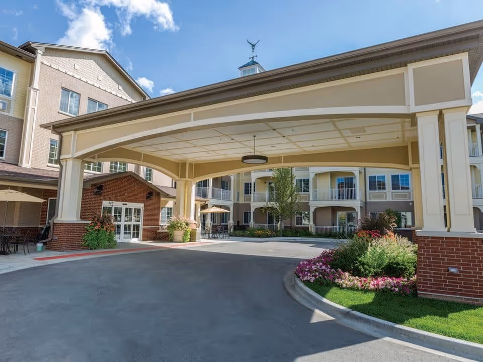 Exterior view of StoryPoint Pickerington senior living facility entrance with a covered driveway, brick and beige building facade, landscaped flower beds, and outdoor seating area under umbrellas.
