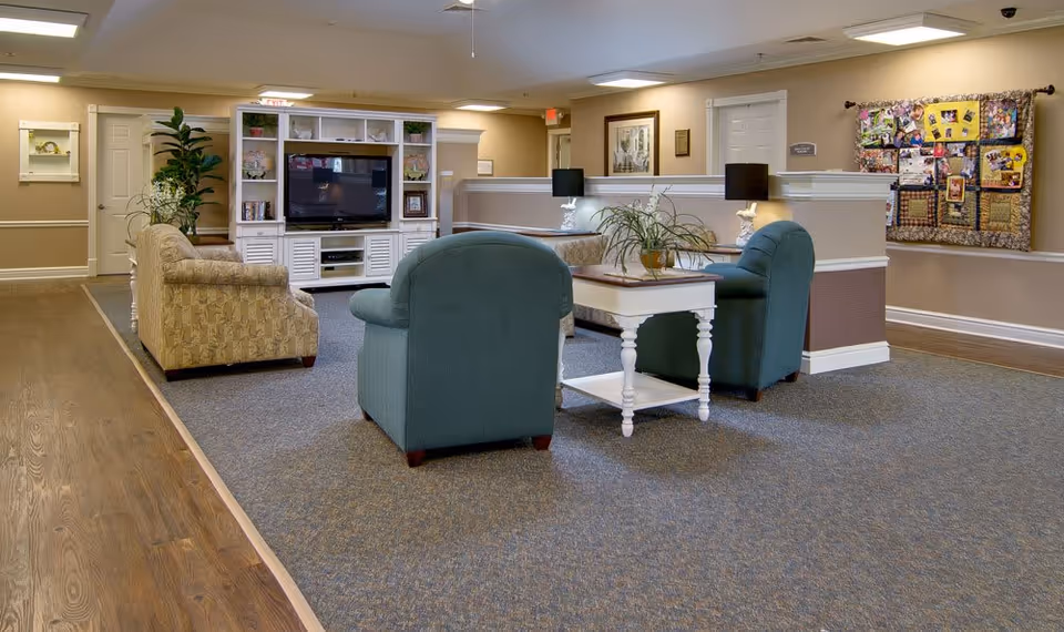 A cozy senior living common area with two green armchairs and one patterned armchair arranged around a white coffee table with a plant on it. A white entertainment center with a TV and decorative items is against the wall. The room has beige walls, carpeted flooring with a wooden border, and a quilted wall hanging on the right side.
