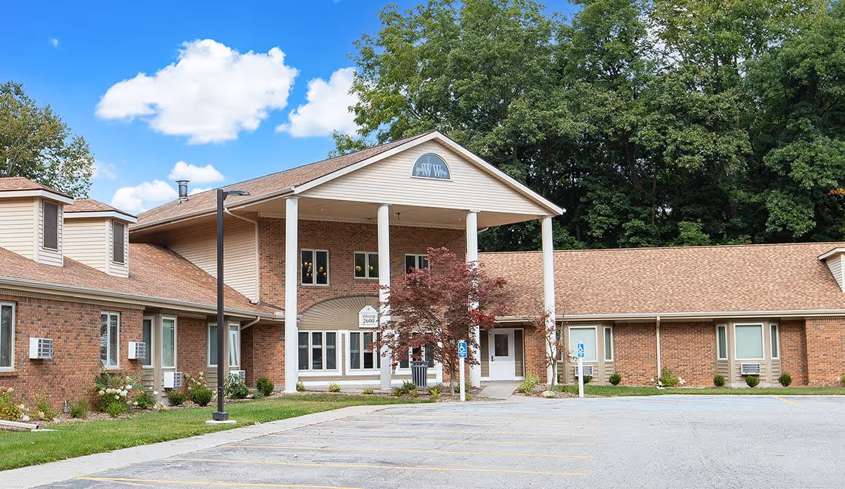 Exterior view of Wyoming Woods facility showing a brick building with a covered entrance supported by four white columns, surrounded by greenery and a parking lot with marked spaces including handicapped spots.