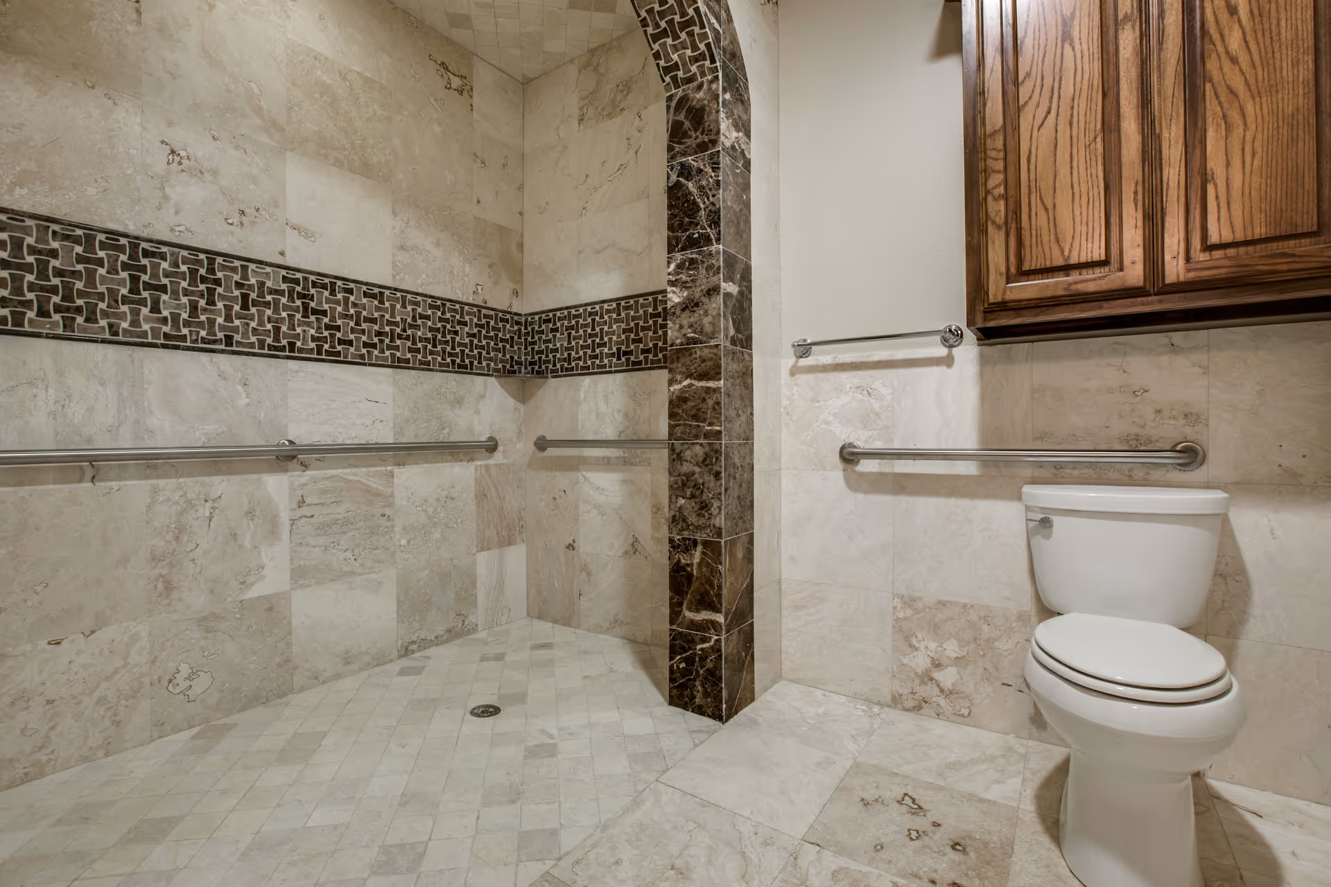 A bathroom with beige marble tiles on the walls and floor, featuring a walk-in shower area with stainless steel grab bars and a decorative dark tile border. There is a white toilet with a stainless steel grab bar next to it and a wooden cabinet mounted on the wall above.