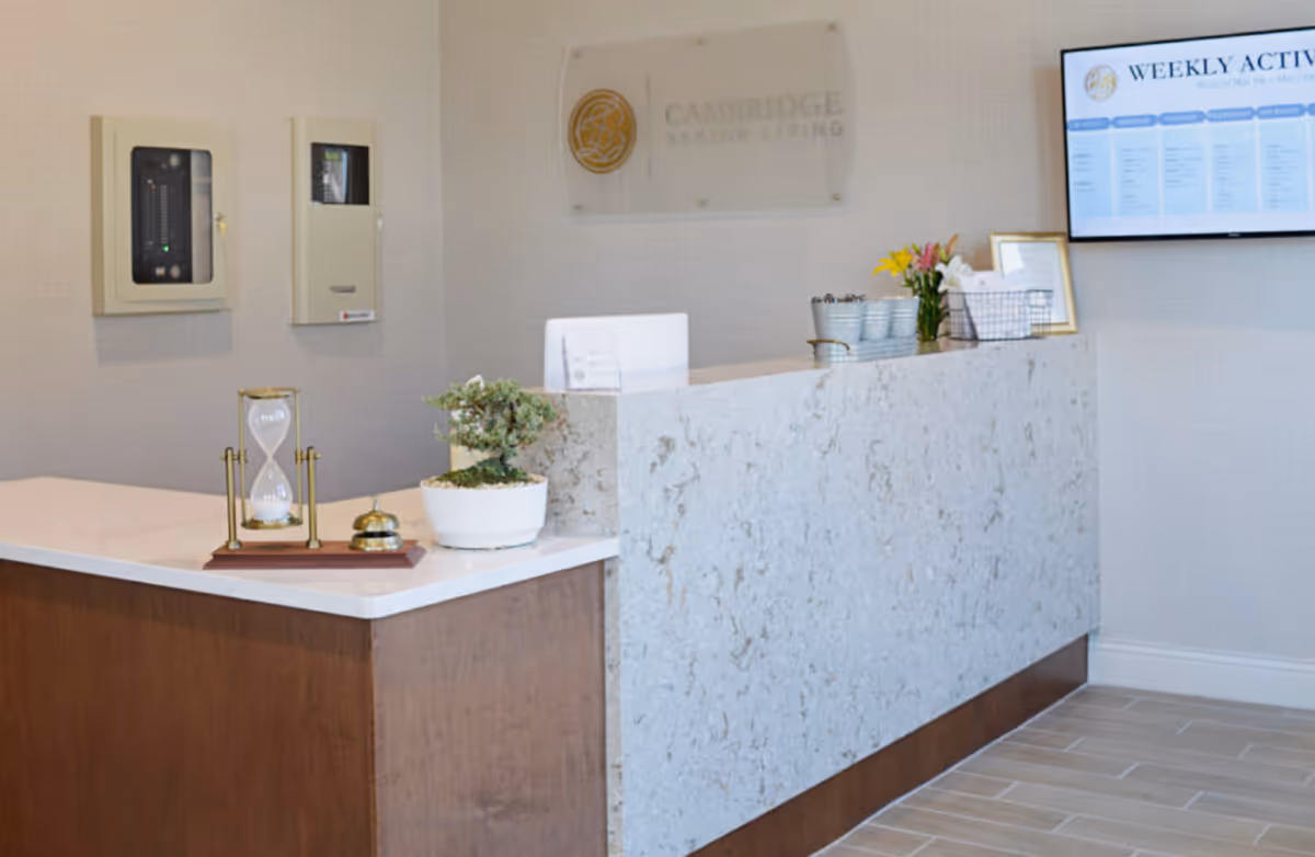 Reception desk area at Cambridge Senior Living with a marble countertop, a small potted plant, an hourglass, a service bell, and a wall-mounted screen displaying weekly activities.