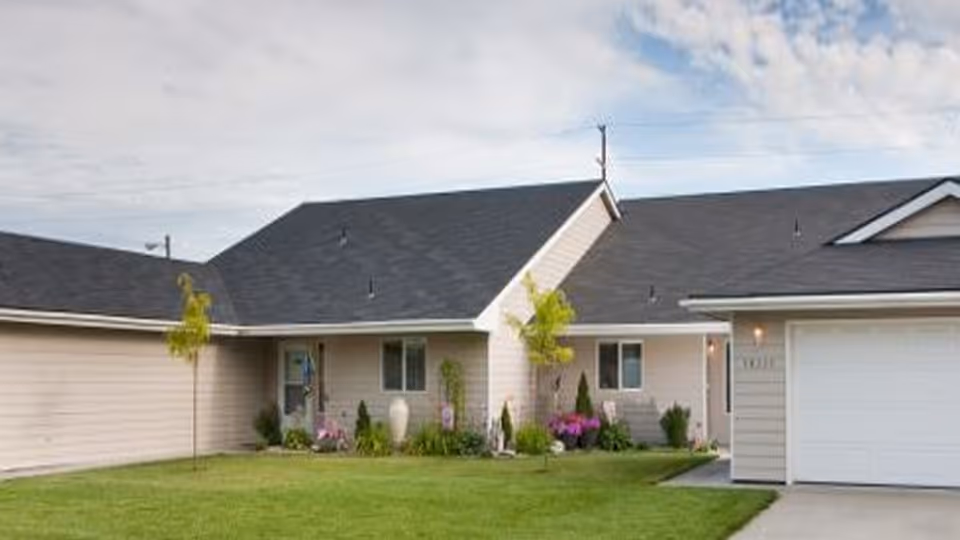 Exterior view of a single-story residential building with beige siding and a dark roof, featuring a well-maintained green lawn, small trees, and flower beds near the entrance and windows.
