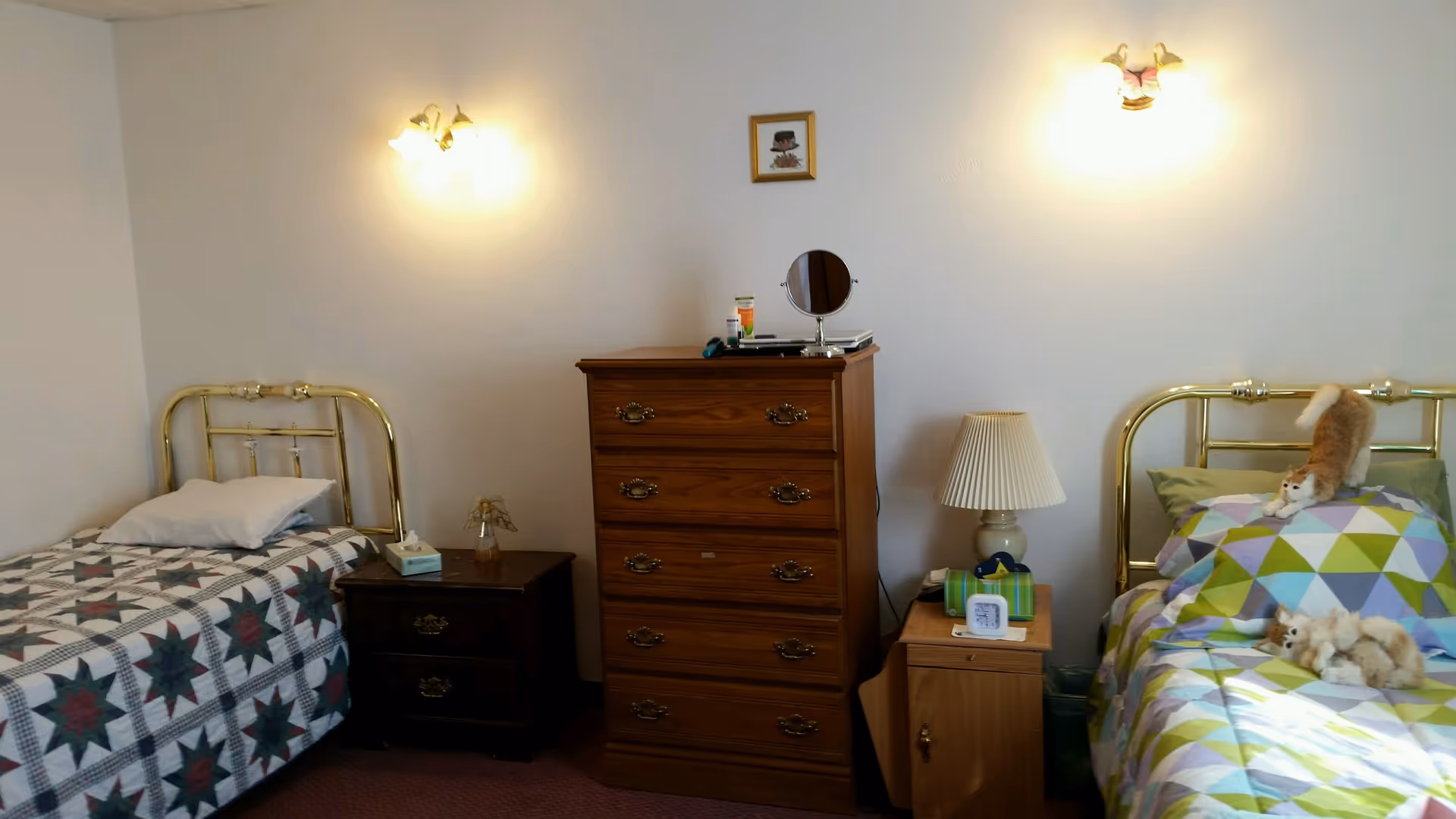 A bedroom with two single beds featuring metal headboards. The bed on the left has a quilt with a star pattern and a white pillow, while the bed on the right has a colorful geometric patterned quilt with two stuffed cats on it. Between the beds is a wooden dresser with four drawers and a small framed picture above it. There are two nightstands, one on each side of the dresser, with a lamp on the right nightstand and a tissue box on the left. The walls are white and there are two wall-mounted light fixtures above each bed.