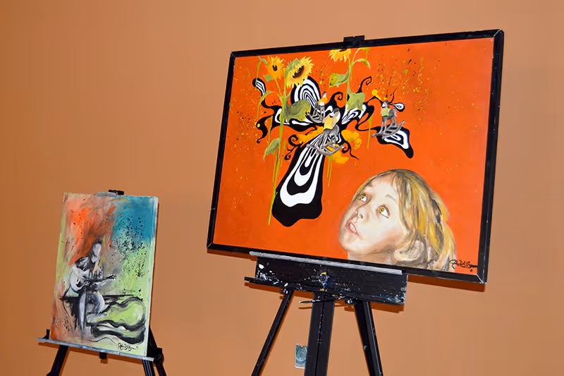 Two paintings displayed on easels against an orange wall. The larger painting features a young girl looking up at abstract black and white shapes intertwined with sunflowers on a bright orange background. The smaller painting depicts a person playing a guitar with a colorful, abstract background.