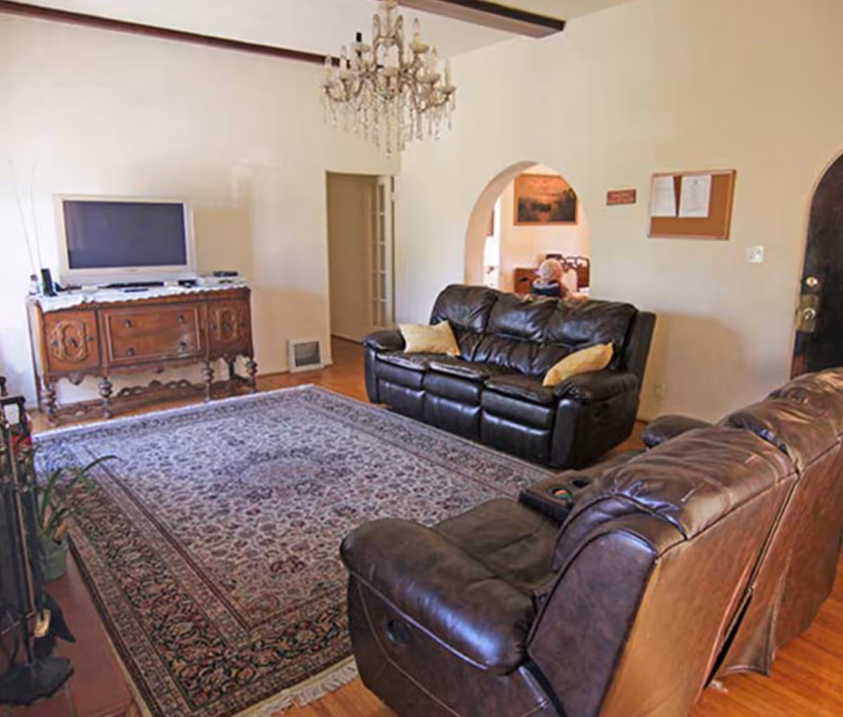 A living room with two dark brown leather sofas, a large patterned area rug, a wooden cabinet with a flat-screen TV on top, a chandelier hanging from the ceiling, and an arched doorway leading to another room.