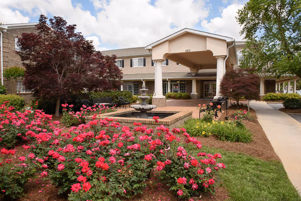 Entrance of The Bridge at Lawrenceville facility with a covered drop-off area, surrounded by a garden featuring vibrant pink flowers, green shrubs, and trees under a partly cloudy sky.